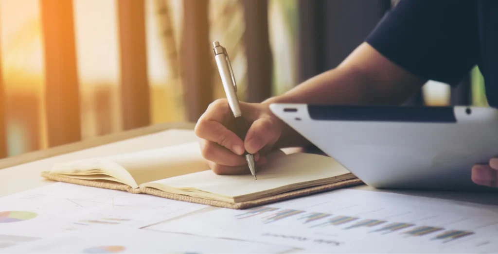 Mains féminines dans un bureau. Une main tient un stylo et écrit dans un carnet, tandis que l'autre tient une tablette.
