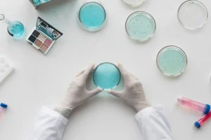 A scientist's hands holding a petri dish. On the bench are research items and a makeup kit, indicating that this is cosmetic research.