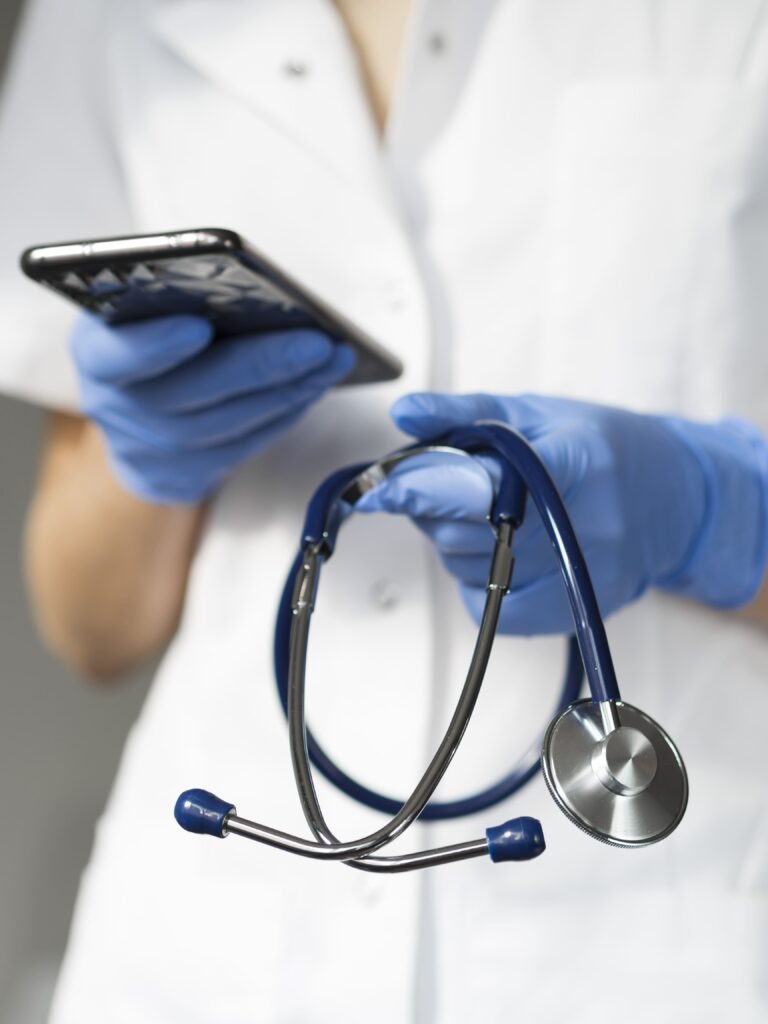 A woman holding a smartphone in one hand and a stethoscope in the other. She wears medical gloves and appears to be in a hospital setting. Regulatory Medical Device Consultancy.