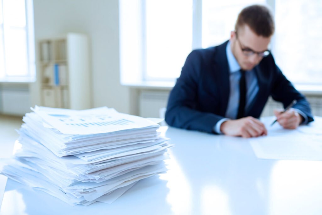 Man at a office desk, looking one document. There is a pile of documents aside him.